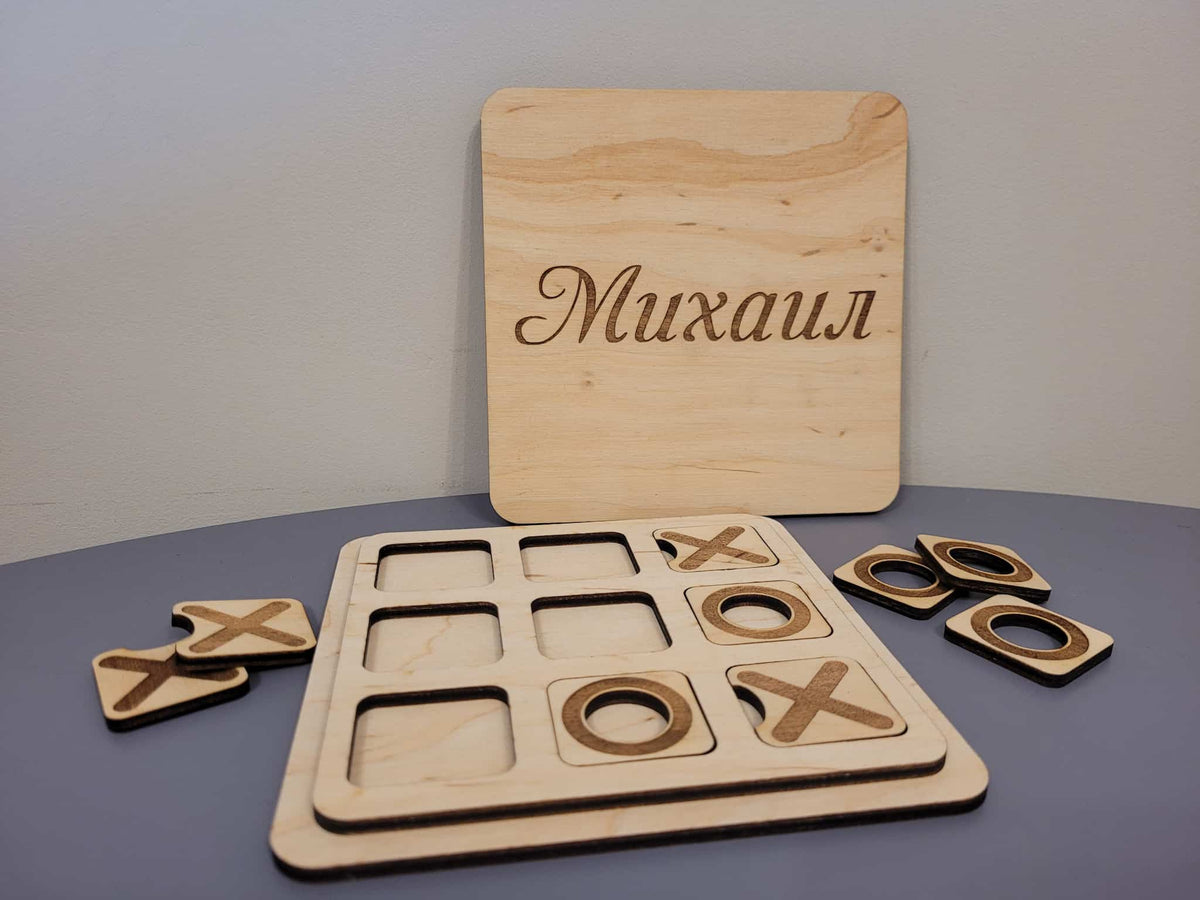 Personalized wooden tic-tac-toe game set with a rectangular board engraved with the name 'Mikhail' in Cyrillic, a game board, and four wooden tokens on a gray surface.