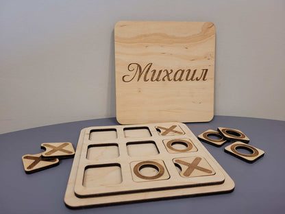 Personalized wooden tic-tac-toe game set with a rectangular board engraved with the name 'Mikhail' in Cyrillic, a game board, and four wooden tokens on a gray surface.