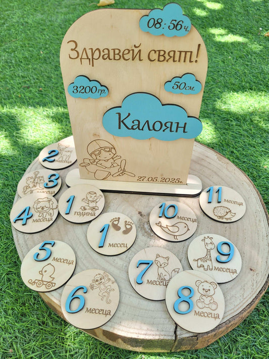 Wooden baby milestone board with engraved numbers, airplane and illustrations on a grassy background.
